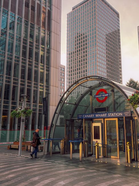 The entrance to Canary Wharf Station featuring a glass and metal curved canopy above the sign, with tall modern office buildings made of glass and steel towering in the background. The scene is illumi