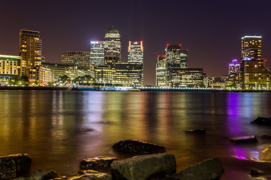 An evening view of a city skyline featuring illuminated modern skyscrapers with glass facades reflecting colorful lights onto the calm river below. The buildings include high-rise offices and commerci