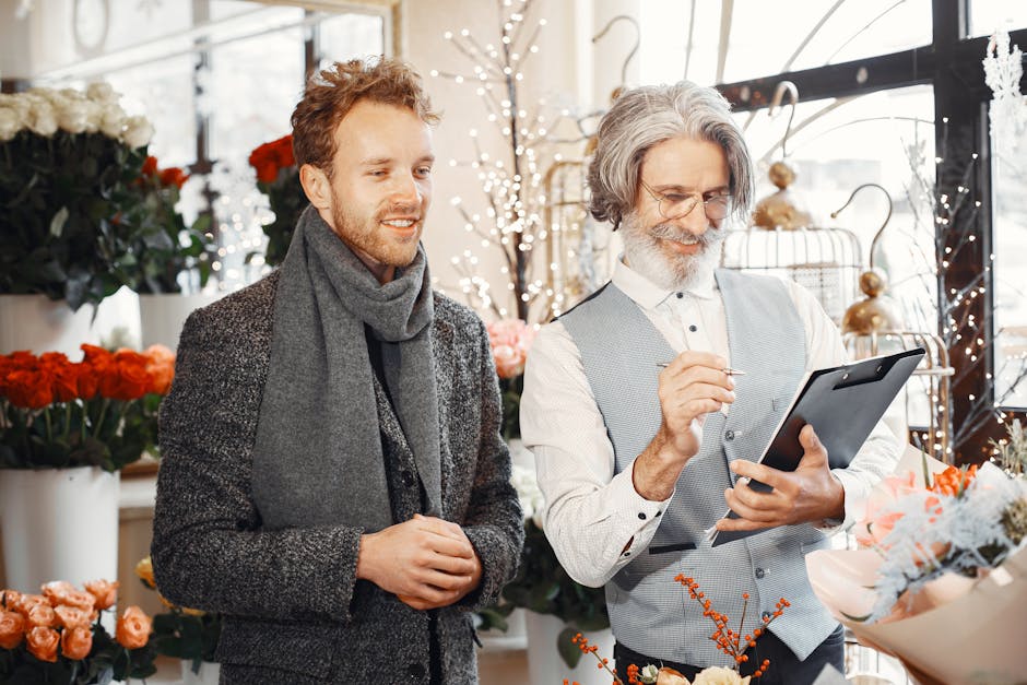 A professional florist with gray hair, glasses, and a white shirt, dressed in a light gray waistcoat, is holding a tablet and pointing at the screen while assisting a young male customer with red hair