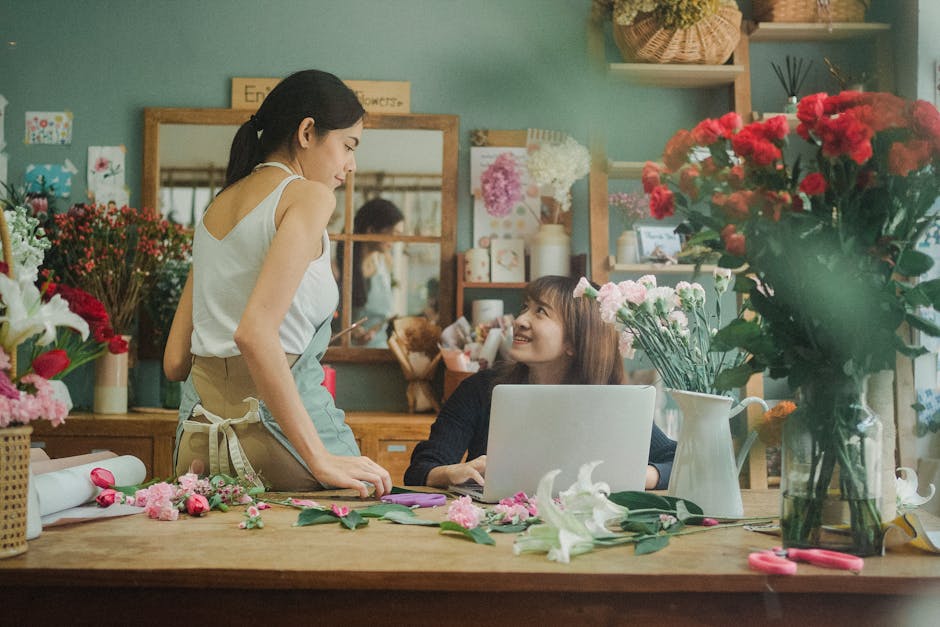 A female florist with dark hair tied back, wearing a white sleeveless top and beige apron, engaging in a floral arrangement in a flower shop. She is leaning slightly forward, holding a pair of scissor