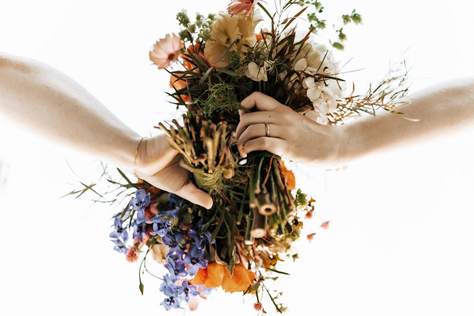 A floral bouquet held by two hands, featuring a mix of fresh flowers including orange poppies, purple delphiniums, white hydrangeas, and green foliage. The arrangement displays a natural, loosely gath