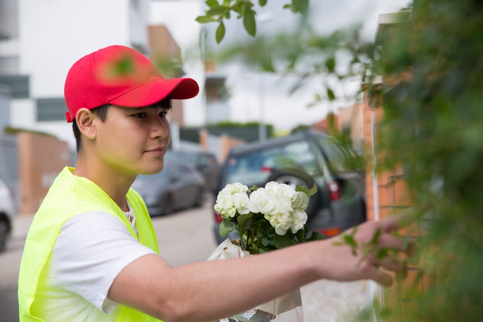 A young male flower delivery courier wearing a red cap and a neon yellow safety vest is placing a fresh bouquet of white roses and hydrangeas against a shrub on a residential street. The arrangement i