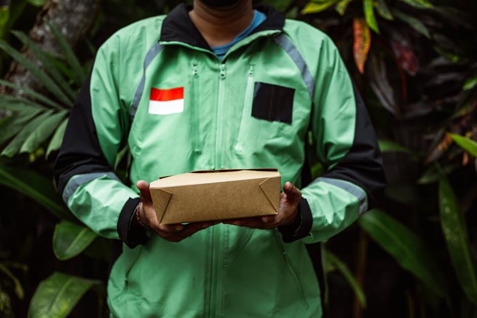 A delivery person wearing a bright green jacket with black accents, standing outdoors against a background of lush, green tropical foliage. The jacket has a small red and white flag patch on the left 