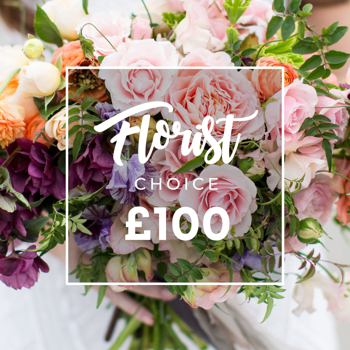 This image captures a richly composed, artisan hand-tied bouquet with a central white rectangular frame reading 'Florist Choice' in flowing script and the clear price '?100' beneath, framed by a delicate white border. The flowers form a layered, garden-style tapestry: large, soft pink garden roses with ruffled, peony-like centers anchor the arrangement and invite touch; clustered low in the design are deep plum and burgundy hellebore-like flowers that lend weight and sophistication. Interspersed are delicate striped sweet peas in pale purple and white, their tendrils suggesting gentle movement, and coral to soft-peach ranunculus providing warm, satiny focal points. Small creamy blooms act as highlights, catching light in tiny glints, while abundant green foliage - from trailing vines to broader textured leaves - builds volume and an effortless, freshly-cut look. The bouquet is held by a pair of hands at the bottom, sleeves blurred in neutral tones so the floral detail remains the star; soft, diffused daylight sculpts petals and shadows, enhancing the tactile quality of each bloom. The overall mood is romantic, refined and hand-crafted - perfect for anniversaries, wedding moments, or as an elevated thank-you. For Millwall residents, the colour story and relaxed structure feel at home beside the docks and green pockets like Millwall Park, a floral voice that mixes retail polish with riverside charm.