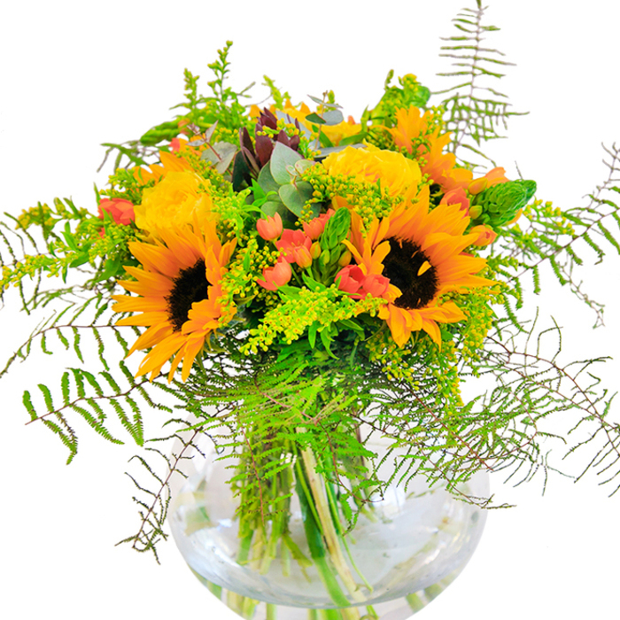 The first impression of this luminous bouquet, crafted near the heart of Millwall, is one of pure exuberance and warmth. Gleaming sunflowers, with their golden petals and inky centers, preside over a vibrant mix of yellow garden roses whose intricate spirals add both visual complexity and timeless charm. Interwoven between the main blooms are lively clusters of bright red Hypericum berries, providing sparkling accents that echo the energy of bustling local markets. Lush, deep green foliage and feathery fern fronds spill outwards, imbuing the design with a sense of movement that feels almost windblown-much like the breezes drifting between the high-rises overlooking the Thames. Light green sprigs of goldenrod pepper the arrangement with tiny, sun-bright blooms, while slender green flower spikes and subtle bluish eucalyptus leaves contribute layers of dimension and gentle aroma. The round, fishbowl-like glass vase elevates the entire composition, revealing crisscrossing stems awash in fresh, clear water-a detail that showcases the freshness and quality local Millwall florists are known for. The bouquet's textures invite the fingertips: suede-soft sunflowers, velvety rose petals, and the crisp snap of greens. With its uplifting palette and expertly balanced design, this bouquet is ideal for festive occasions or as a radiant gesture of gratitude delivered across SE16. It embodies the best of seasonal floristry and the welcoming spirit of Millwall's close-knit community.