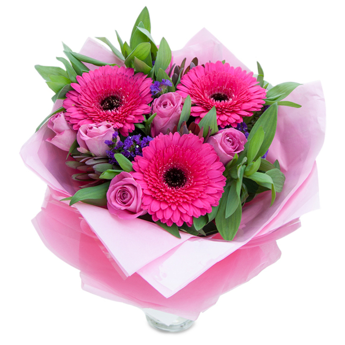 I always favour bouquets that balance boldness with a whisper of softness; this composition does exactly that with its vivid fuchsia and muted lavender-pink tones. Three exuberant Gerbera daisies punch through the arrangement with saturated fuchsia petals, their dark centres providing a compelling visual contrast and a rhythmic focal point. Surrounding them, roses in a dusty lavender-pink sit in their early stages of bloom, petals folded in graceful spirals that feel both comforting and romantic. Interspersed sprigs of purple statice lend a wildflower touch-tiny starry clusters that break up the larger shapes and introduce a pleasing, papery texture. The foliage, including broad, glossy leaves reminiscent of salal and the soft grey-green of eucalyptus, creates a lush, garden-like envelope that gives the bouquet weight and a sense of fullness. The bouquet is meticulously wrapped: an inner sheet of matte pale pink tissue provides a soft base while an outer layer of translucent fuchsia-pink cellophane, slightly crinkled, adds depth and a polished sheen; the folds are deliberate and neat, a sign of careful florist craft. The clear glass vase rim shows at the bottom, suggesting immediacy and fresh water for the stems. Under soft morning light the velvety petals reveal subtle gradients and the scent profile is gentle-roses offering a soft perfume and gerberas a light, green-sweet hint. This arrangement feels intimate and uplifting, perfect for placing on a kitchen table or gifting to mark a special moment in Millwall, near Manchester Road DLR or the cafés by Canary Wharf.